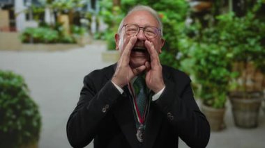 Takım elbiseli kıdemli bir adam, arka planda yeşillik olan bir parkta tutkulu bir şekilde konuşuyor. Başarı ya da rekabeti gösteren bir madalya takıyor..