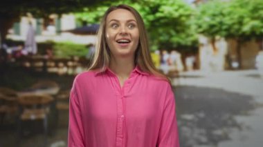 Güneşli bir şehir caddesinde, bulanık binalar ve ağaçlarla gülümseyen bir kadın, şaşkın, neşeli bir ifadeyle kentsel anı yakalıyor..