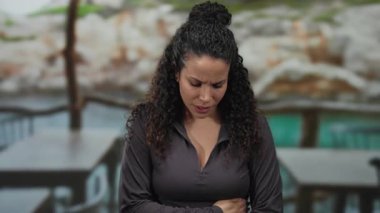 Kıvırcık saçlı bir kadın sahilde karnını tutarak ayakta duruyor, iyi hissetmiyor gibi görünüyor, deniz kenarında bir çevresi olduğunu düşündüren kayalık bir geçmişi var..