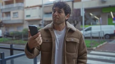 İspanyol bir adam, şehir manzarası ve binalarla çevrili bir şekilde telefona gülümserken sokakta selfie çekiyor. Sıradan bir ceket giyiyor ve etrafa mutluluk ve güven saçıyor..