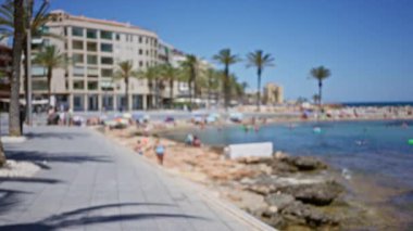 Bulanık insanlar sahilde güneşli bir günün tadını çıkarıyorlar. Arka planda yüksek binalar ve palmiye ağaçları var.