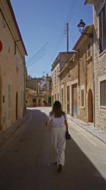 Kadın, Mallorca 'da eski bir Akdeniz kasabasında, taşralı taş binalar ve açık mavi gökyüzü ile çevrili güneşli dar bir sokakta geziniyor..