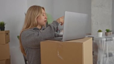 Yeni bir evde dizüstü bilgisayar üzerinde çalışan bir kadın. Etrafı kutular ve bitkilerle çevrili. Rahat bir atmosferi olan rahat ve modern bir iç mekan sergiliyor..