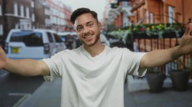 Sakallı genç adam, kolları açık, beyaz gömlekli, şehir mimarisi ve park halindeki araçlara karşı açık havada gülümsüyor..