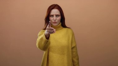 Sarı bir süveteri basit bir kahverengi duvar arka planına doğrultup kafa karışıklığını ifade eden bir kadın, kırmızı saçlarına ve günlük tarzına odaklanmış duygu jestleri sergiliyor.