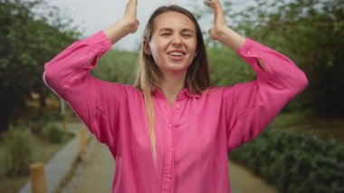 Pembe tişörtlü genç bir kadın, güneşli bir günde iki eliyle başını tutarak yeşil bir parkta boğulmuş hissediyor. Duygularını ve doğasını gösteriyor..