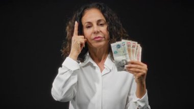 Beyaz gömlekli bir kadın, elinde İngiliz sterlini banknotlarıyla siyah arka plan karşısında kameraya ciddi bir ifadeyle bakıyor ve para kavramını vurguluyor..