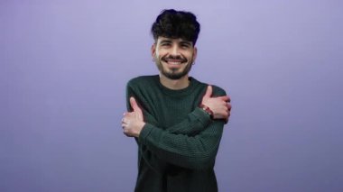 Kollarını göğsüne dolayıp gülümseyen, mor arka planda yeşil kazak giyen, izole ortamda mutluluk ve güven saçan genç bir adam.