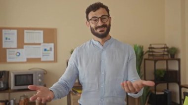 Sakallı İspanyol bir adam modern bir ofiste elinde tablolarıyla gülümsüyor ve el kol hareketi yapıyor. Sıcak ve misafirperver bir ortam sunuyor..