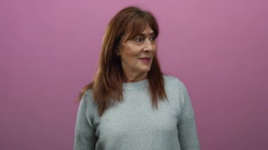 Üst düzey kadın pembe arka planda gülümsüyor, süveter giyiyor, neşeli bir ifade sergiliyor ve stüdyo ortamında mutluluk iyimserliğini ve sıcaklığını iletiyor..