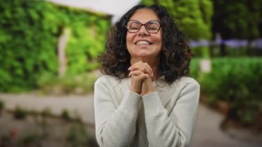 Parkta gülümseyen bir kadın, gözlüklü ve süveterli, yemyeşil, mutluluk ve sükuneti somutlaştıran.