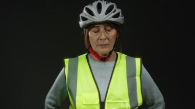 Üst düzey İspanyol kadın, siyah arka plana karşı duruyor. Kaskı ve yansıtıcı yeleği var. Güvenli ve izole edilmiş bir stüdyoya odaklanıyor..