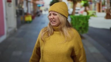 İğrendiğini ifade eden sarı kazak ve bereyle şehir caddesinde duran bulanık geçmişli bir kadın.