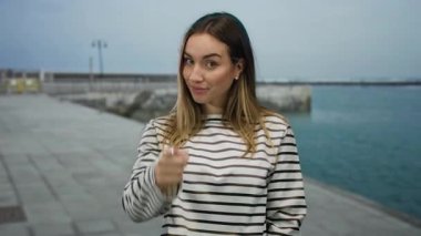 Deniz kenarında gezinen, çekici ifadeleri, sarı saçları ve çizgili gömleği olan, bulutlu gökyüzünün altında neşeli bir ruh hali sergileyen bir kadın..