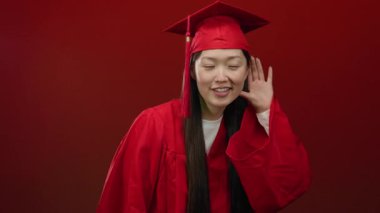 Kırmızı mezuniyet elbisesi giymiş, gülümseyen bir kadın canlı kırmızı bir arka plana karşı bir jest yaparak coşkusunu ve heyecanını gösteriyor..