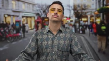 Genç İspanyol bir adam kentin hareketli caddesinde kendine güveni tam bir şekilde duruyor. Şık güneş gözlüğü ve şehir hayatı dolu desenli bir gömlek giyiyor..