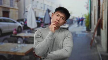 Genç bir adam açık hava restoranının terasında dikilirken düşünceli bir ifadeyle bulanık bir şehir geçmişine karşı.