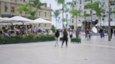 Bulanık yayalar Monako 'da geziyor. Etrafı lüks binalar ve kafelerle çevrili. Palmiye ağaçlarıyla dolu kalabalık bir sokak ortamında..