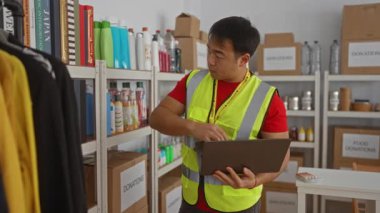 Parlak yelekli genç adam bağış merkezinde raflarda eşyalar düzenliyor, elinde dijital tabletle verimli depolama çözümlerine odaklanıyor..