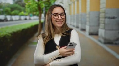 Şehir sokağında elinde telefonla gülümseyen, gözlük takan, günlük giysiler giyen, yürüyüş yolunun yanında duran, arkasında canlı yapraklar olan kadın rahat ve mutlu görünüyor..