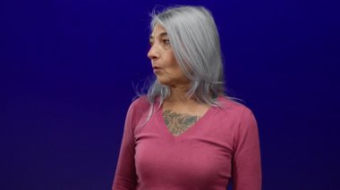 Pembe tişörtlü yaşlı, gri saçlı bir kadın izole edilmiş mavi arka plana şaşırmış görünüyor, samimi bir duygusal ifade yakalıyor..