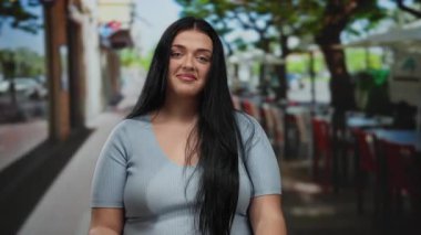 Restoranın terasında uzun saçlı, iri yarı genç bir kadın masaların ve yeşilliklerin bulanık arka planına sahip sıradan bir ortamda kendine güvenini ve tarzını sergiliyor..