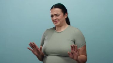 Mavi bir arkaplana karşı iğrenme hareketi yapan açık yeşil gömlek giyen, negatif duygu ve vücut dilini basit bir stüdyo ortamında sergileyen bir kadın..