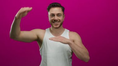Sakallı genç adam canlı pembe arka plana karşı duruyor beyaz atlet giyiyor ve gülümsüyor, pozitifliğini ve enerjisini gösteriyor..