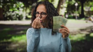 Parkta elinde Arjantin pezosu olan, gözlüklü ve kazaklı orta yaşlı bir kadın ağaçlara ve arka planda güneş ışığına olan öfkesini dile getiriyor..