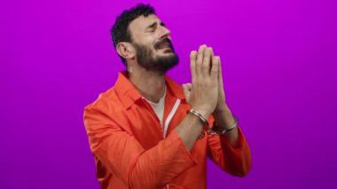 Elleri kelepçeli, turuncu tulumlu İspanyol bir adam canlı, mor bir arka plana karşı duruyor..