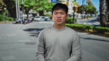 Sokaktaki genç adam mutsuz görünüyor. Arka planda yeşillik ve araçlar var. Şehir hayatının bir anını yakalıyor..