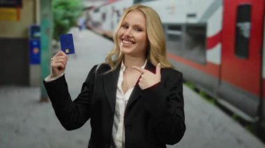 Tren istasyonunda elinde kredi kartıyla gülümseyen, arka planda trenleri olan siyah ceket ve beyaz gömlek giyen bir kadın, seyahat ve finansmanı öneriyor..