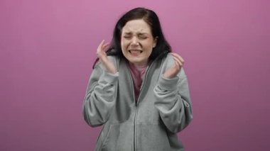 Gri kapüşonlu bir kadın canlı pembe bir arka plana karşı anlamlı bir yüzle poz veriyor, günlük tarzını ve esmer saçlarını sergiliyor..