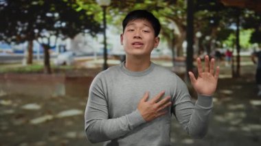 Genç Çinli bir adam, sakin bir açık hava parkında, gün ışığının altında el hareketleriyle duygularını ifade ederek iletişim için barışçıl ve çekici bir atmosfer yaratıyor..
