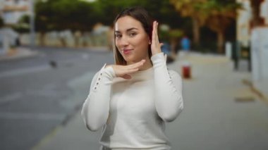 Bir sokak ortamında coşkuyla el kol hareketi yapan bir kadın genç enerjisini ve tarzını canlı şehir arkaplanına karşı sergiliyor sarı saçlı ve beyaz bluzlu.