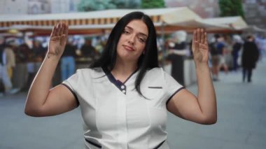 Elleri açık şehir pazarında poz veren kadın, insanların alışveriş ve hareketli market tezgahlarının arka planına karışıyor..