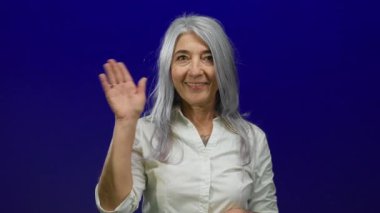 Gri saçlı, gülümseyen ve kameraya el sallayan yaşlı bir kadın. Mavi, izole bir arka plan duvarına karşı. Dostane ve davetkar bir jest sergiliyor..