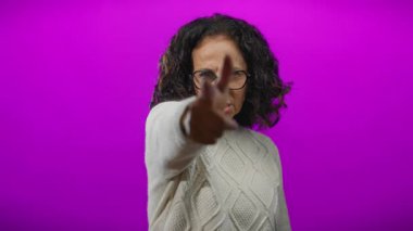 Kıvırcık saçlı olgun bir kadın, canlı mor bir arka plana karşı neşeli bir parmak tabancası hareketi yapar. Kendine güven ve neşe ifade eder..