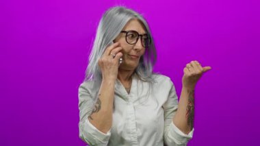 Gri saçlı yaşlı kadın akıllı telefondan konuşuyor ve pembe arka planda baş parmağıyla hareketler yapıyor, gözlük takıyor ve kolunda dövmeleri olan beyaz bir gömlek giyiyor..