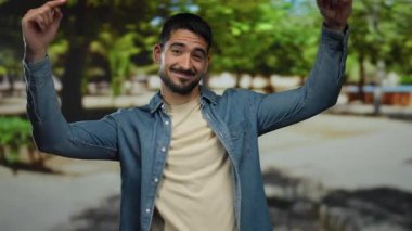 Kot gömlekli genç adam güneşli bir park ortamında gülümsüyor ve onu işaret ediyor, ağaçlı ve rahat bir atmosferi olan sıradan bir doğa yaşam tarzını sergiliyor..