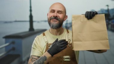 Sakallı, eldivenli kel bir adam. Arka planda bulutlu bir gökyüzü olan deniz kenarında kese kağıdı tutuyor..