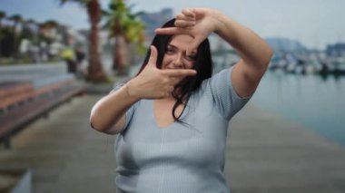 Kadın deniz kenarındaki bir gezinti alanında yaratıcı bir şekilde el kol hareketi yapıyor. Kaygısız, kıvrımlı tarzını sahilde neşeli bir ifadeyle kucaklıyor. Etrafı sakin bir arka planla çevrili..