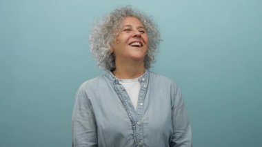 Gri saçlı bir kadın, izole edilmiş mavi bir duvara sevinçle gülüyor. Rahat bir poz vererek mutluluk ve kendine güven yayıyor. Orta yaşlı zarafetini sergiliyor..