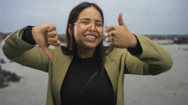 Deniz kenarındaki kumsalda, bulutlu gökyüzünün altında, mizahi bir jest yaparak baş parmaklarını yukarı aşağı gösteren genç bir kadın.