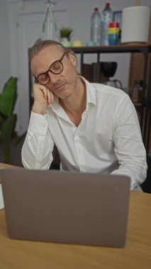 Ofiste gözlüklü, beyaz tişörtlü, dizüstü bilgisayarda çalışan orta yaşlı beyaz bir adam yorgun görünüyor ve arka planda rafları olan bir masada oturuyor..