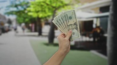 Bulanık şehir manzarası arka planında ABD dolarlarını elinde tutan bir adam, açık şehir ortamında Amerikan para birimini gözler önüne seriyor, finansal konsepti vurguluyor..