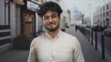 Sakallı genç adam canlı bir şehir caddesinde duruyor, gözlük takıyor ve açık tişört giyiyor, rahat ve şehir dışı yaşam tarzını yansıtıyor..