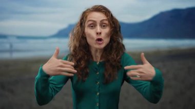 Sahilde enerjik bir şekilde el kol hareketi yapan, arka planda dağlar olan bir kadın, açık deniz kenarında iletişim ve ifadeye vurgu yapıyor..