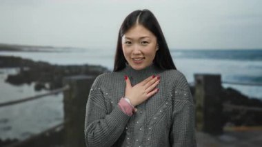 Deniz kenarından hoşlanan uzun saçlı bir kadın kumsala bakıyor, doğal güzelliğini ve huzurlu atmosferini sergiliyor..