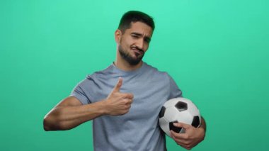 Elinde futbol topu tutan genç adam, kendinden emin ve çekici poziyonuyla yeşil duvar arkasında olumlu ve sportmence durduğunu gösteriyor..